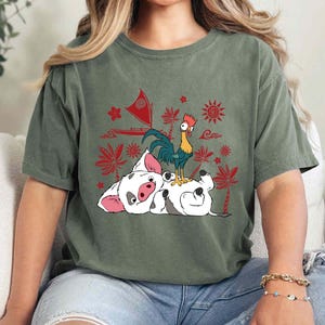 May include: Olive green t-shirt featuring a graphic of a white pig and a rooster with red and yellow accents. The pig is lying down, and the rooster is standing on top of it. Red floral and sun designs are in the background.