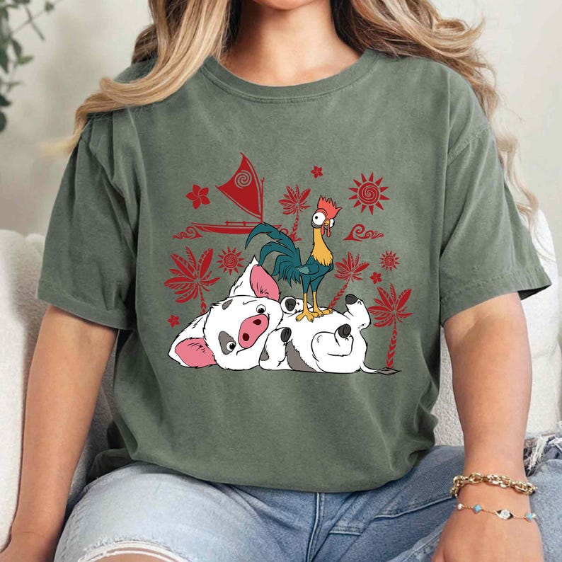 May include: Olive green t-shirt featuring a graphic of a white pig and a rooster with red and yellow accents. The pig is lying down, and the rooster is standing on top of it. Red floral and sun designs are in the background.