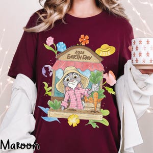 Op de afbeelding: Bordeauxrood T-shirt met een cartoon konijn in een tuinscene, met wortels en de tekst "2026 Earth Day". Het konijn draagt een roze geruit overhemd en een strohoed. Kleurrijke bloemen en een klein paars konijn silhouet zijn ook aanwezig.