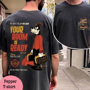 May include: Pepper-colored t-shirt featuring a cartoon bellhop character in a red uniform, holding luggage. The shirt has the text "YOUR ROOM IS READY" and "THE HOLLYWOOD TOWER HOTEL" printed on it. The shirt is a dark gray color.
