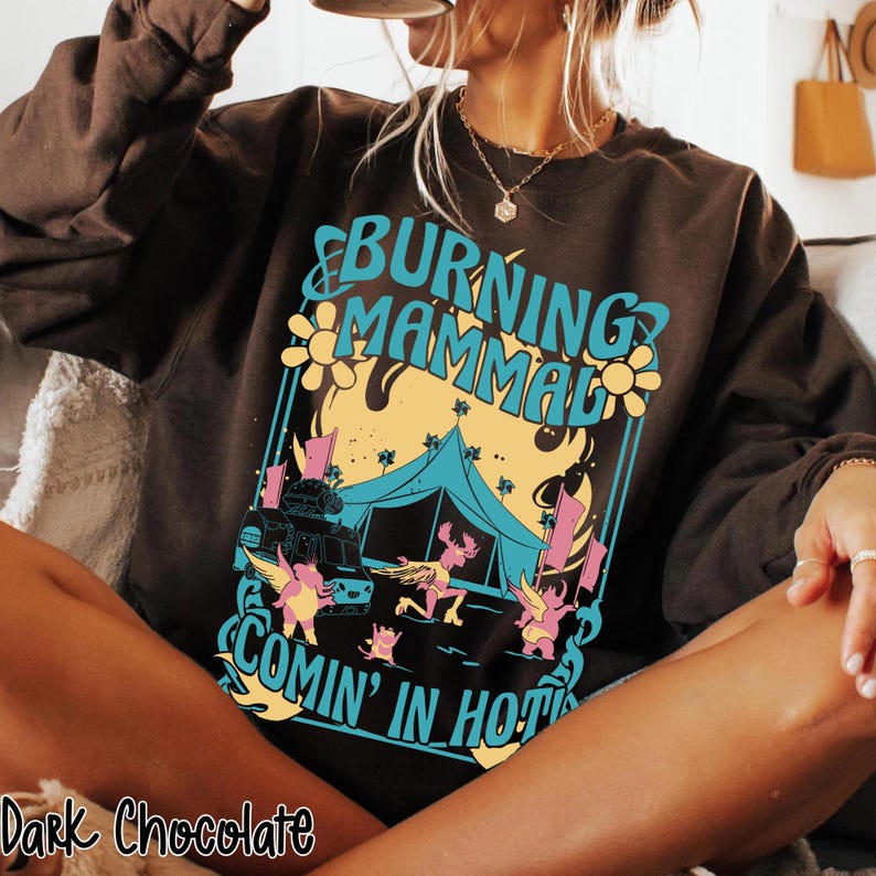 Puede incluir: Sudadera de color chocolate oscuro con un dise&ntilde;o gr&aacute;fico. El dise&ntilde;o presenta el texto "BURNING MAMMAL" en turquesa, una llama amarilla y naranja, una tienda de campa&ntilde;a azul y figuras de dibujos animados. El texto "COMIN' IN HOT" est&aacute; debajo.