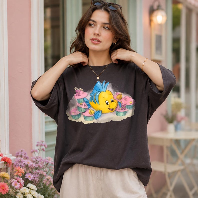 May include: Dark gray t-shirt featuring a cartoon image of a yellow fish with blue fins and a smiling face, surrounded by pink cupcakes. The shirt has short sleeves and a relaxed fit, suitable for casual wear.