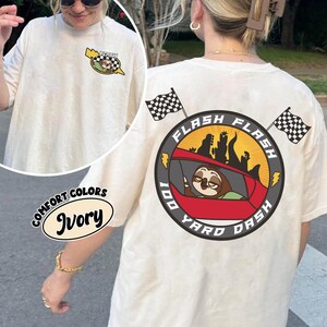 May include: Ivory-colored t-shirt with a graphic on the front and back. The back graphic features a sloth in a red car with the words "Flash 100 Yard Dash." The front graphic has a sloth in a car with the word "Flash."