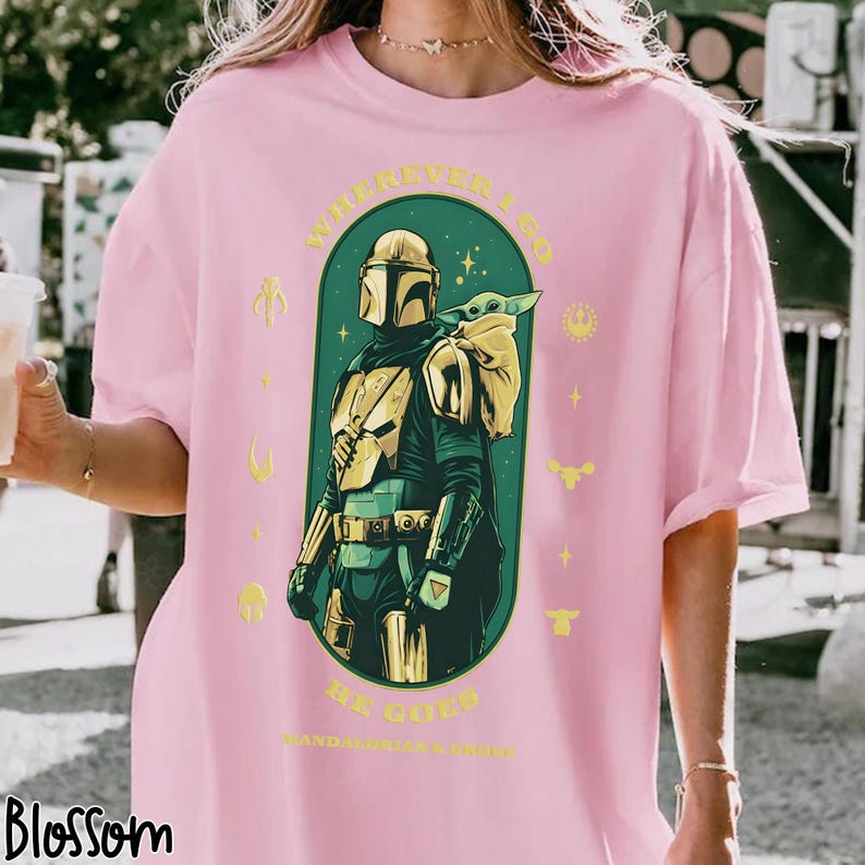 Peut inclure: T-shirt rose clair avec un motif de The Mandalorian et Grogu. Le design comprend le texte "WHENEVER I GO, BE GOOD, MANDALORIAN'S GUIDE" en jaune. L'illustration est dans un ovale vert avec des &eacute;toiles et des accents jaunes.