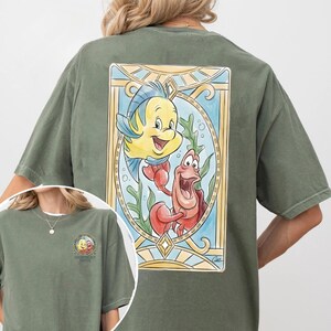 May include: Olive green t-shirt featuring a colorful illustration of Flounder and Sebastian from The Little Mermaid. The design is framed with a stained-glass style border. A smaller version of the design is on the front.