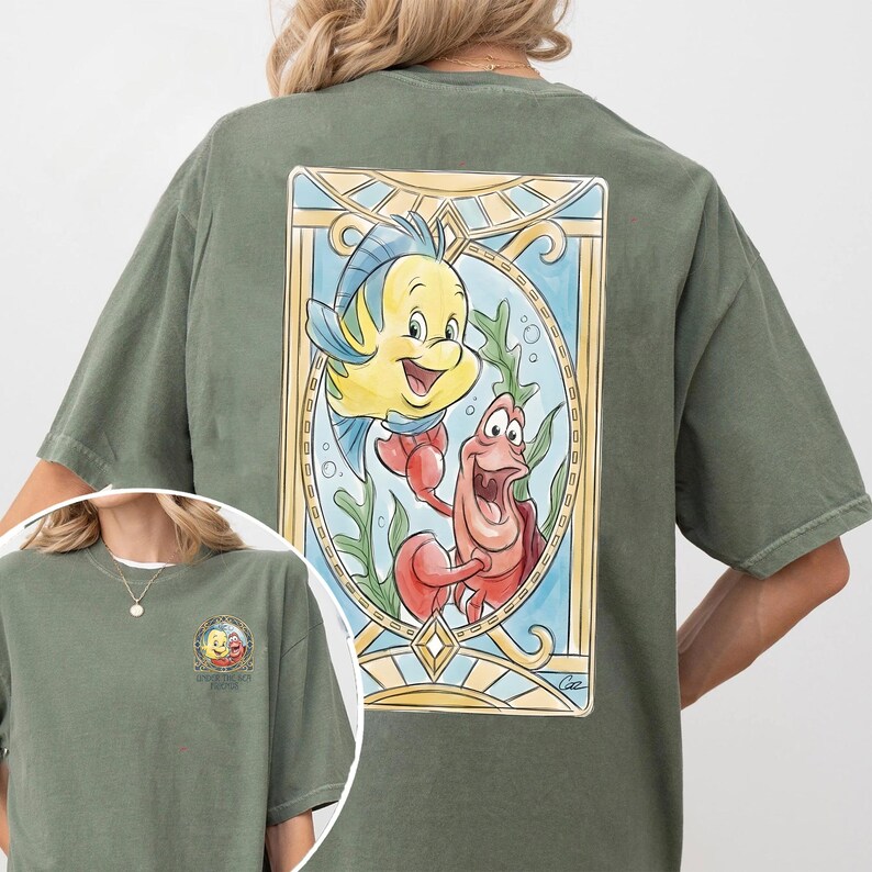 May include: Olive green t-shirt featuring a colorful illustration of Flounder and Sebastian from The Little Mermaid. The design is framed with a stained-glass style border. A smaller version of the design is on the front.