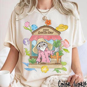Op de afbeelding: Cr&egrave;mekleurig T-shirt met een cartoonkonijn in een roze geruit overhemd en strohoed, met wortels en groenten. Het ontwerp bevat een marktkraam met de tekst "2026 Earth Day", bloemen en een wereldbol. Er is ook een gele hoed met "2026".