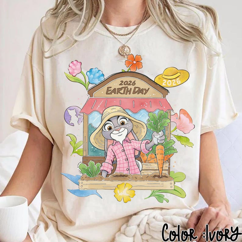 Op de afbeelding: Cr&egrave;mekleurig T-shirt met een cartoonkonijn in een roze geruit overhemd en strohoed, met wortels en groenten. Het ontwerp bevat een marktkraam met de tekst "2026 Earth Day", bloemen en een wereldbol. Er is ook een gele hoed met "2026".