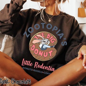 Puede incluir: Sudadera de color chocolate oscuro con el texto "Zootopia's The Big Donut" y una imagen de dibujos animados de Judy Hopps. Debajo del gr&aacute;fico de la rosquilla est&aacute; el texto "Little Rodentia".