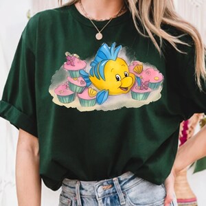 May include: Dark green t-shirt featuring a cartoon illustration of a yellow and blue fish character surrounded by pink cupcakes. The fish has a smiling expression. The cupcakes have pink frosting and are in light blue cupcake liners.