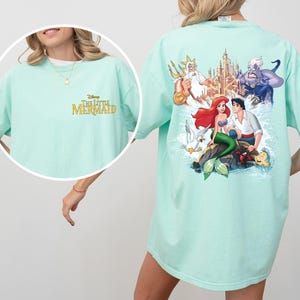 May include: A light blue t-shirt with a graphic print of characters from the Disney movie The Little Mermaid. The text "The Little Mermaid" is printed in gold on the front of the shirt.