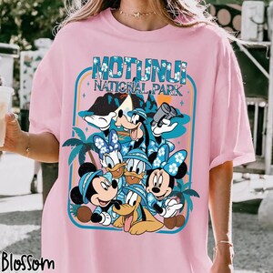 May include: Pink t-shirt featuring a graphic of Disney characters, including Mickey, Minnie, Donald, Daisy, Goofy, and Pluto, with the text "Motunui National Park." The design is framed with a blue border and palm trees.