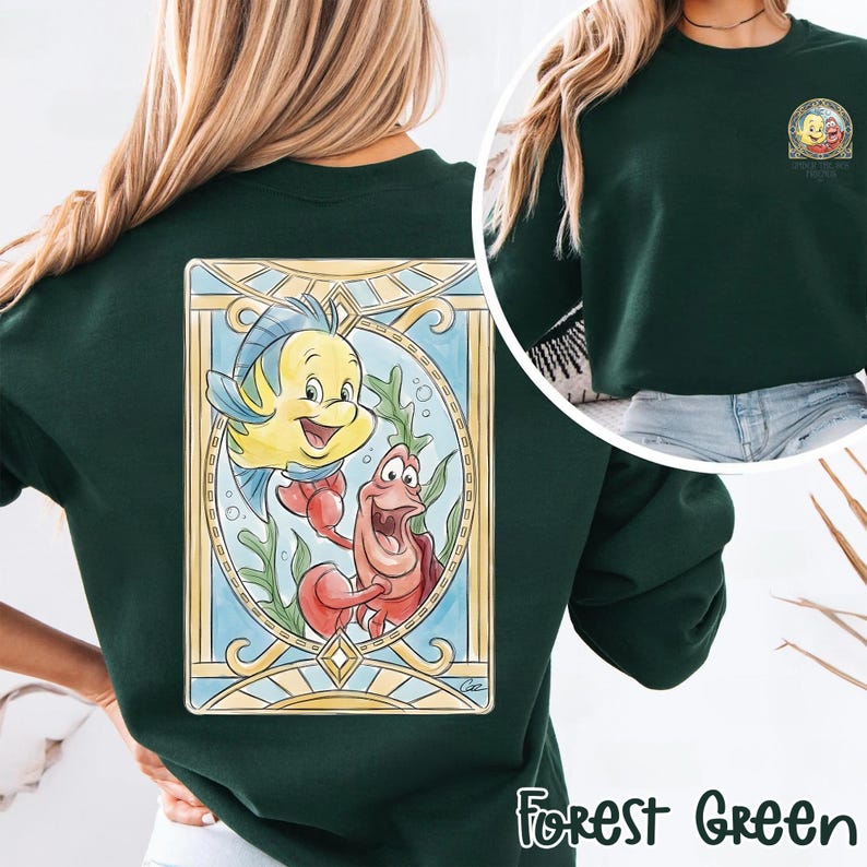 May include: Forest green sweatshirt featuring a colorful illustration of Flounder and Sebastian from The Little Mermaid. The design is framed in a stained-glass style, with the words "Forest Green" printed at the bottom.