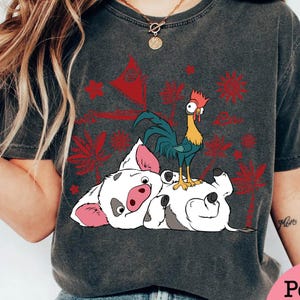 May include: Dark gray t-shirt featuring a cartoon illustration of a pig lying on its back with a rooster standing on top. Red and white tropical design elements are printed on the shirt.