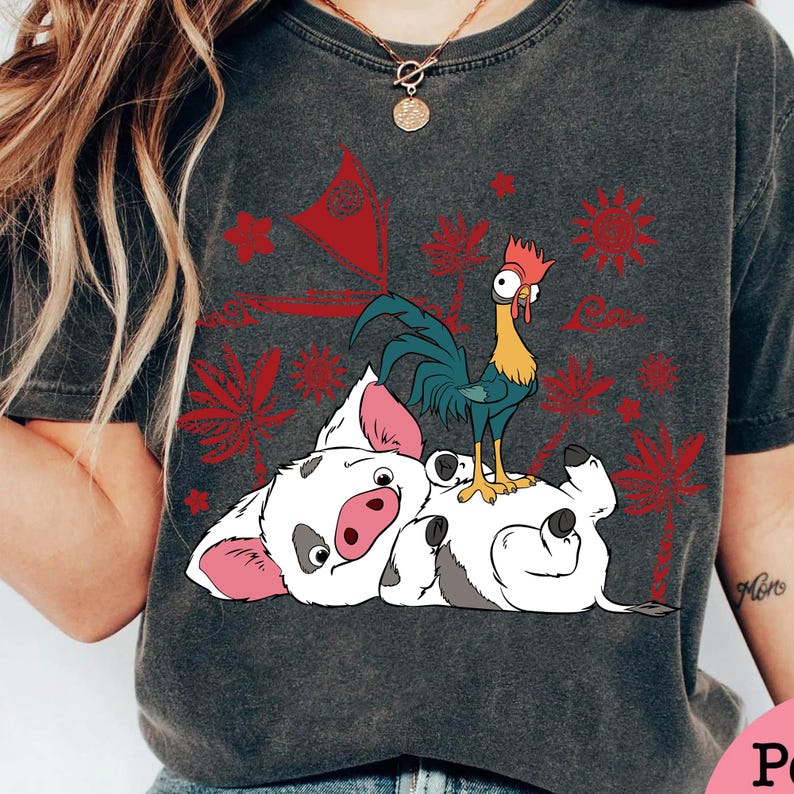 May include: Dark gray t-shirt featuring a cartoon illustration of a pig lying on its back with a rooster standing on top. Red and white tropical design elements are printed on the shirt.