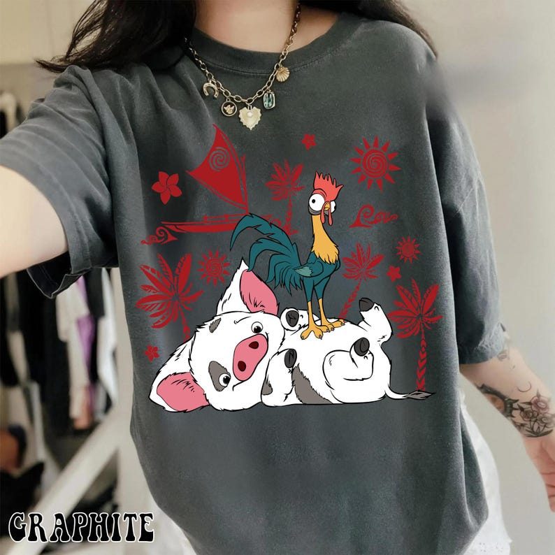 May include: Graphite-colored t-shirt featuring a cartoon illustration of a pig and a rooster. The pig is lying on its back, and the rooster is standing on top of it. Red floral and sun designs are in the background.