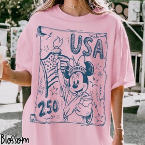 May include: Pink t-shirt with a blue graphic featuring Minnie Mouse as the Statue of Liberty, the American flag, the word "USA," and the number "250." The design is in a vintage style.