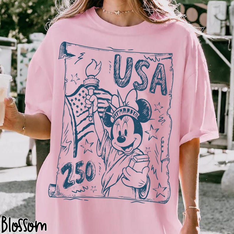 May include: Pink t-shirt with a blue graphic featuring Minnie Mouse as the Statue of Liberty, the American flag, the word "USA," and the number "250." The design is in a vintage style.