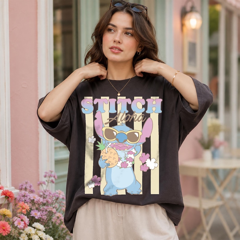 K&ouml;nnte beinhalten: Dunkelgraues T-Shirt mit der Figur Stitch aus Lilo & Stitch, die eine Sonnenbrille tr&auml;gt und eine Ananas h&auml;lt. Der Text "Stitch Aloha" ist in Pastellfarben gehalten, mit vertikalen Streifen an den Seiten.