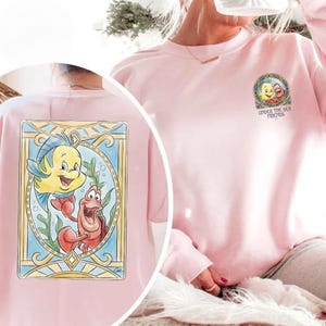 May include: Light pink sweatshirt featuring a colorful graphic of Flounder and Sebastian from The Little Mermaid. The design is framed in a stained-glass style. The text "UNDER THE SEA FRIENDS" is also visible.