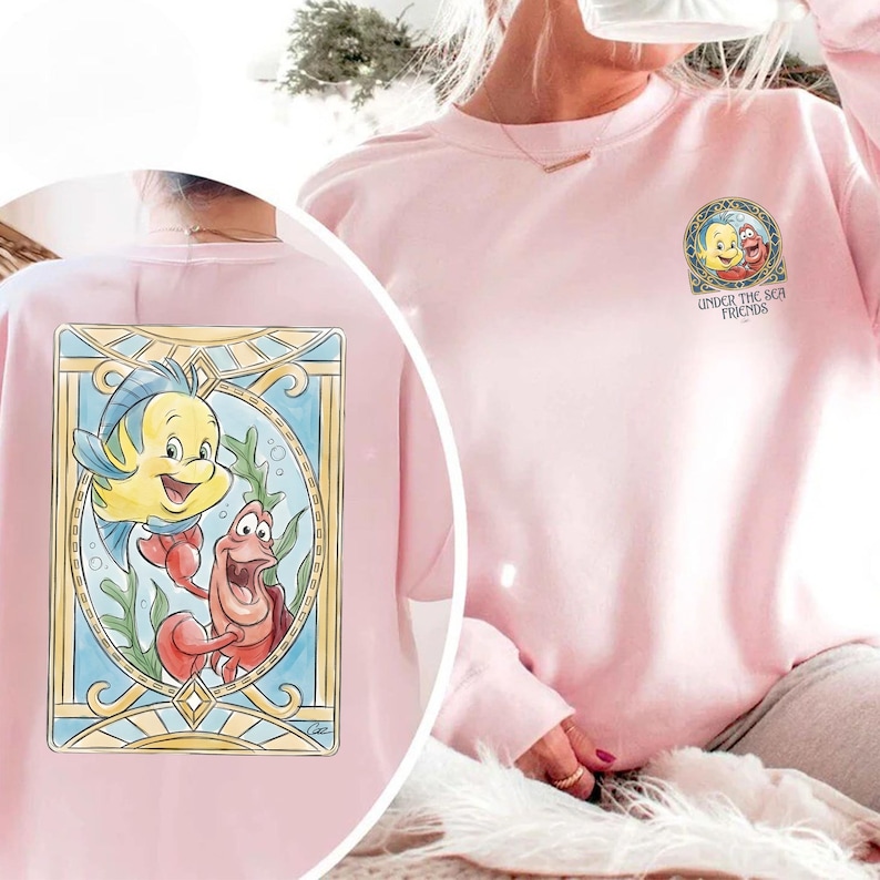 May include: Light pink sweatshirt featuring a colorful graphic of Flounder and Sebastian from The Little Mermaid. The design is framed in a stained-glass style. The text "UNDER THE SEA FRIENDS" is also visible.