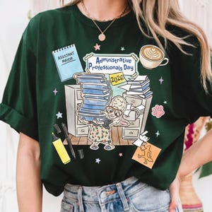 Puede incluir: Camiseta verde oscuro con una ilustraci&oacute;n caprichosa que celebra el D&iacute;a de los Profesionales Administrativos 2026. El dise&ntilde;o presenta una oveja de dibujos animados que lleva libros, una taza de caf&eacute; y material de oficina. El texto incluye "Assistant Mayor".