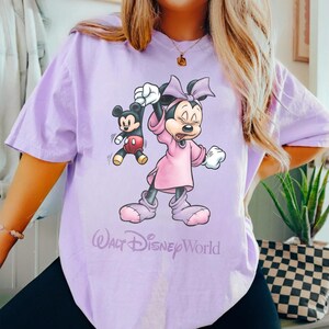 May include: A light purple t-shirt featuring a graphic of Minnie Mouse holding Mickey Mouse. Minnie is wearing a pink dress and bow. The text "Walt Disney World" is printed below the graphic.