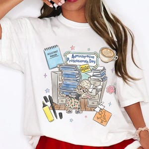 Puede incluir: Camiseta blanca con una colorida ilustraci&oacute;n para el D&iacute;a de los Profesionales Administrativos 2026. El dise&ntilde;o incluye una pila de libros, un personaje de conejo, caf&eacute; y el texto "Assistant Mayor".