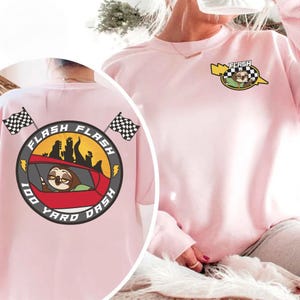 May include: A light pink sweatshirt with a cartoon sloth design. The front features a small graphic with a sloth in a race car. The back has a larger design with the text "FLASH 100 YARD DASH" and checkered flags.