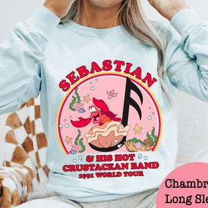 May include: Light blue long-sleeve shirt featuring a graphic of Sebastian the crab from The Little Mermaid. The graphic includes the text "Sebastian & His Hot Crustacean Band 1991 World Tour" in red and pink. The shirt is a chambray long sleeve.
