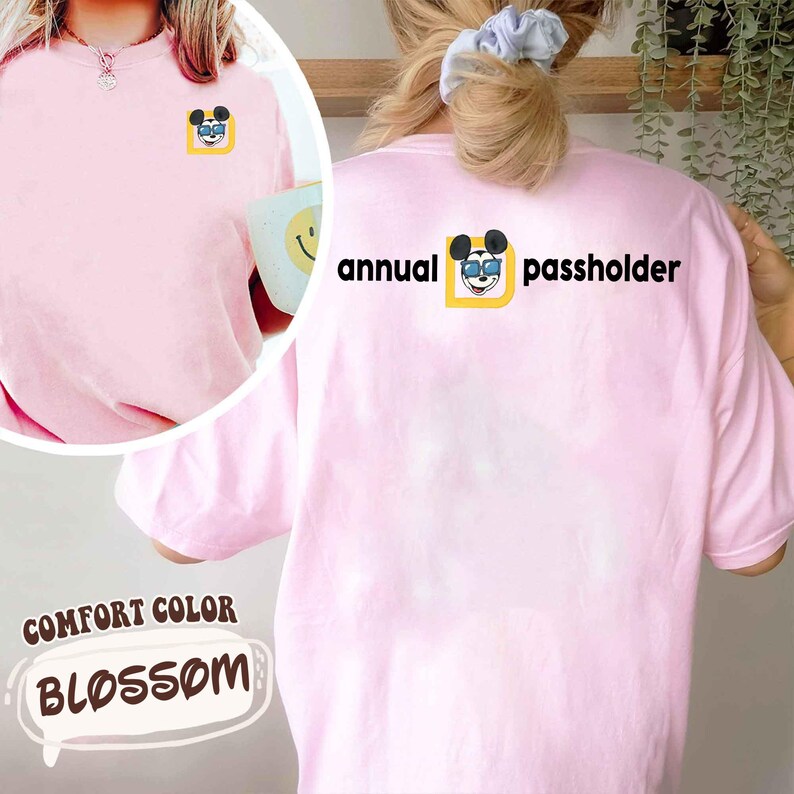 Puede incluir: Una camiseta rosa claro Comfort Colors Blossom con un peque&ntilde;o gr&aacute;fico de Mickey Mouse en la parte delantera y un gr&aacute;fico m&aacute;s grande en la parte trasera. El gr&aacute;fico de la espalda dice "annual passholder" en texto negro. La camiseta es de corte holgado.