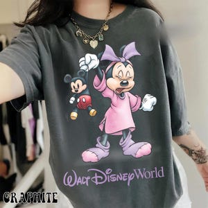May include: Graphite-colored t-shirt featuring a graphic of Minnie Mouse holding Mickey Mouse. Minnie is wearing a pink dress and bow. The text "Walt Disney World" is printed in purple. The word "GRAPHITE" is printed at the bottom.