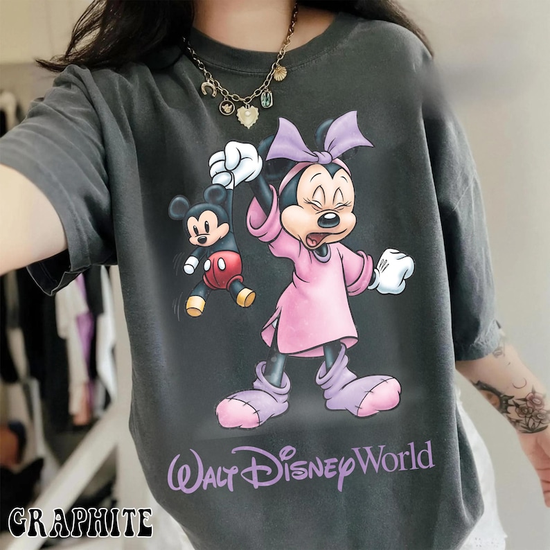 May include: Graphite-colored t-shirt featuring a graphic of Minnie Mouse holding Mickey Mouse. Minnie is wearing a pink dress and bow. The text "Walt Disney World" is printed in purple. The word "GRAPHITE" is printed at the bottom.