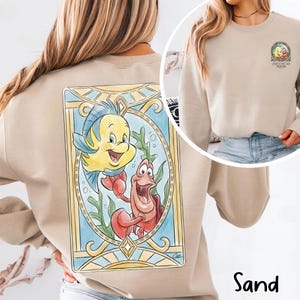 May include: Sand-colored sweatshirt featuring a colorful illustration of Flounder and Sebastian from The Little Mermaid. The design is framed in a stained-glass style with a blue and gold border. The sweatshirt also has a small graphic on the front.