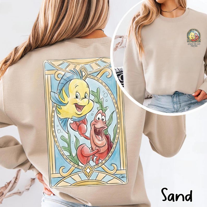 May include: Sand-colored sweatshirt featuring a colorful illustration of Flounder and Sebastian from The Little Mermaid. The design is framed in a stained-glass style with a blue and gold border. The sweatshirt also has a small graphic on the front.