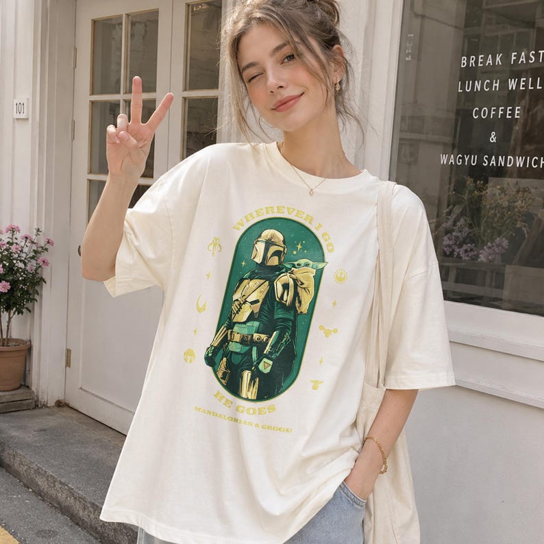 Peut inclure: T-shirt cr&egrave;me avec un motif de The Mandalorian et Grogu. Le design est encadr&eacute; dans un ovale vert et comprend le texte "WHEREVER I GO HE GOES". Le t-shirt a des manches courtes et une coupe ample.