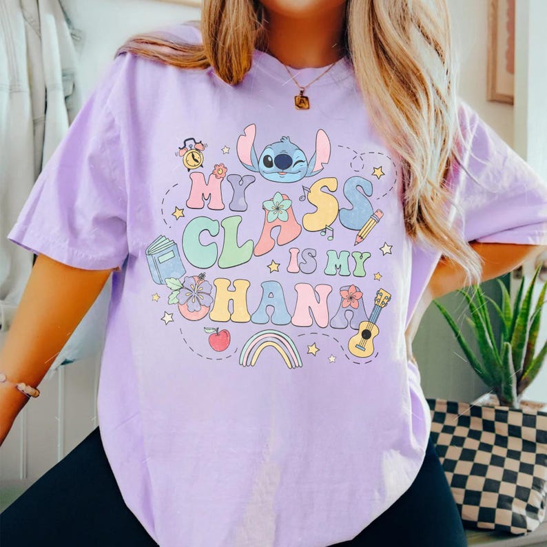 Puede incluir: Camiseta de color lila claro con la frase "My Class is My Ohana" en letras coloridas de estilo caricatura. El dise&ntilde;o incluye a Stitch, un arco&iacute;ris, estrellas e ilustraciones escolares.
