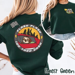 May include: Forest green sweatshirt featuring a cartoon sloth driving a red car with the text "FLASH 100 YARD DASH". The design includes a checkered flag and a yellow lightning bolt. The sweatshirt has a crew neck.
