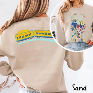 Pu&ograve; includere: Felpa color sabbia con una grafica colorata di un treno sul retro e un design di personaggi dei cartoni animati sul davanti. Il design frontale include il testo "We're Anyone, We Can Be Anything."