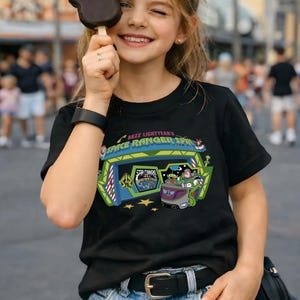 K&ouml;nnte beinhalten: Schwarzes T-Shirt mit einer farbenfrohen Grafik von Buzz Lightyear's Space Ranger Spin. Das Design zeigt den Eingang der Attraktion, Buzz Lightyear und andere Charaktere. Der Text "Buzz Lightyear's Space Ranger Spin" ist ebenfalls zu sehen.
