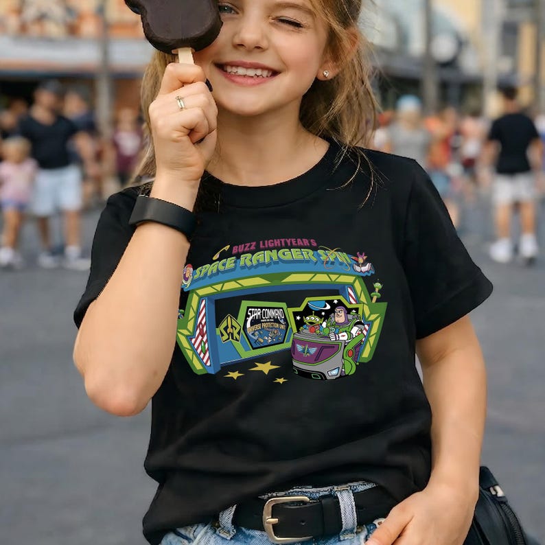 K&ouml;nnte beinhalten: Schwarzes T-Shirt mit einer farbenfrohen Grafik von Buzz Lightyear's Space Ranger Spin. Das Design zeigt den Eingang der Attraktion, Buzz Lightyear und andere Charaktere. Der Text "Buzz Lightyear's Space Ranger Spin" ist ebenfalls zu sehen.