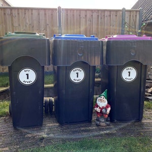 Op de afbeelding: Drie zwarte recyclebakken met verschillende gekleurde deksels. De bakken zijn gelabeld met witte stickers waarop "GFT 1 Afval", "Papier 1 Afval" en "Rest 1 Afval" staat. Een kleine tuinkabouter staat voor de bakken.