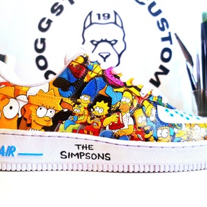 Puede incluir: Zapatillas blancas con un diseño personalizado que presenta personajes de Los Simpson. Las zapatillas tienen un logotipo "AIR" azul y el texto "THE SIMPSONS" en el lateral.