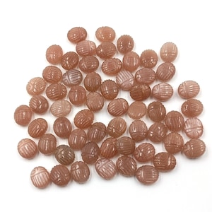May include: A collection of carved, oval-shaped beads in shades of brown and peach. Each bead features a detailed, textured design. The beads are scattered on a white surface, creating a visually appealing arrangement. These beads are suitable for jewelry making.