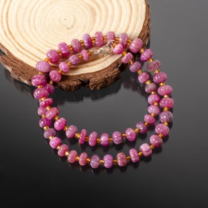 Op de afbeelding: Een roze robijn edelsteen ketting met een zilveren sluiting. De ketting is gemaakt van kleine, ronde kralen en wordt getoond op een houten oppervlak.