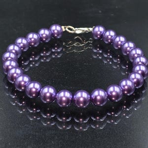 May include: A circular bracelet composed of uniform, smooth, deep purple spherical beads. The beads are strung together and secured with a silver-toned metal lobster claw clasp, creating a continuous loop of polished, dark violet circular elements.