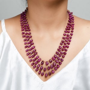 May include: A multi-strand necklace made of teardrop-shaped, deep purple gemstones. The necklace is made of gold-toned metal.