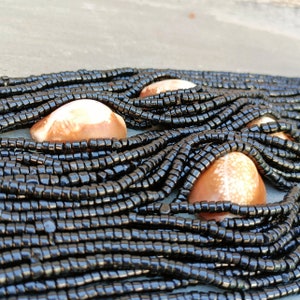 K&ouml;nnte beinhalten: Eine schwarze Perlenkette mit braunen Muscheln. Die Kette besteht aus vielen Str&auml;ngen kleiner schwarzer Perlen und hat mehrere gro&szlig;e, glatte Muscheln.