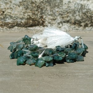 May include: A strand of green agate gemstone beads in a star shape. The beads are strung on silver wire and have a white tassel.