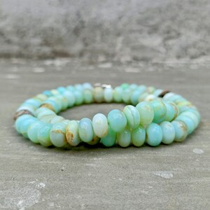 May include: A necklace made of light blue and white oval-shaped beads. The beads are strung together, forming a circular shape. The necklace is resting on a gray surface.